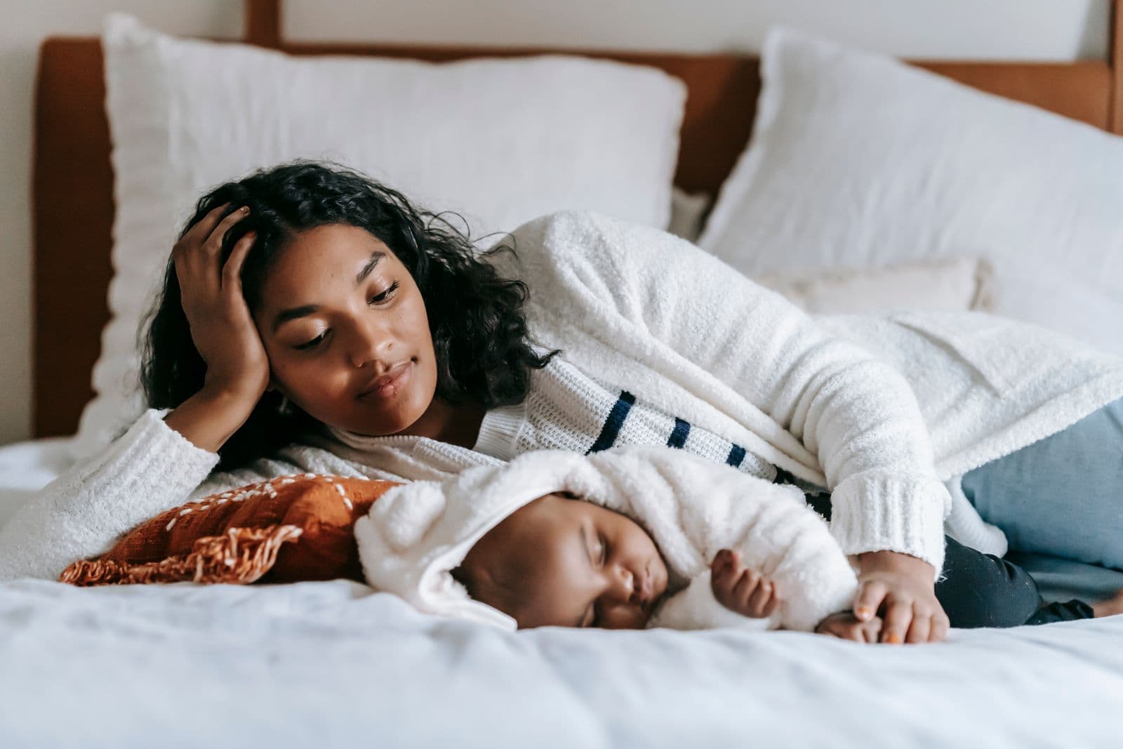A mother lies beside her sleeping baby on a white bed in morning light.