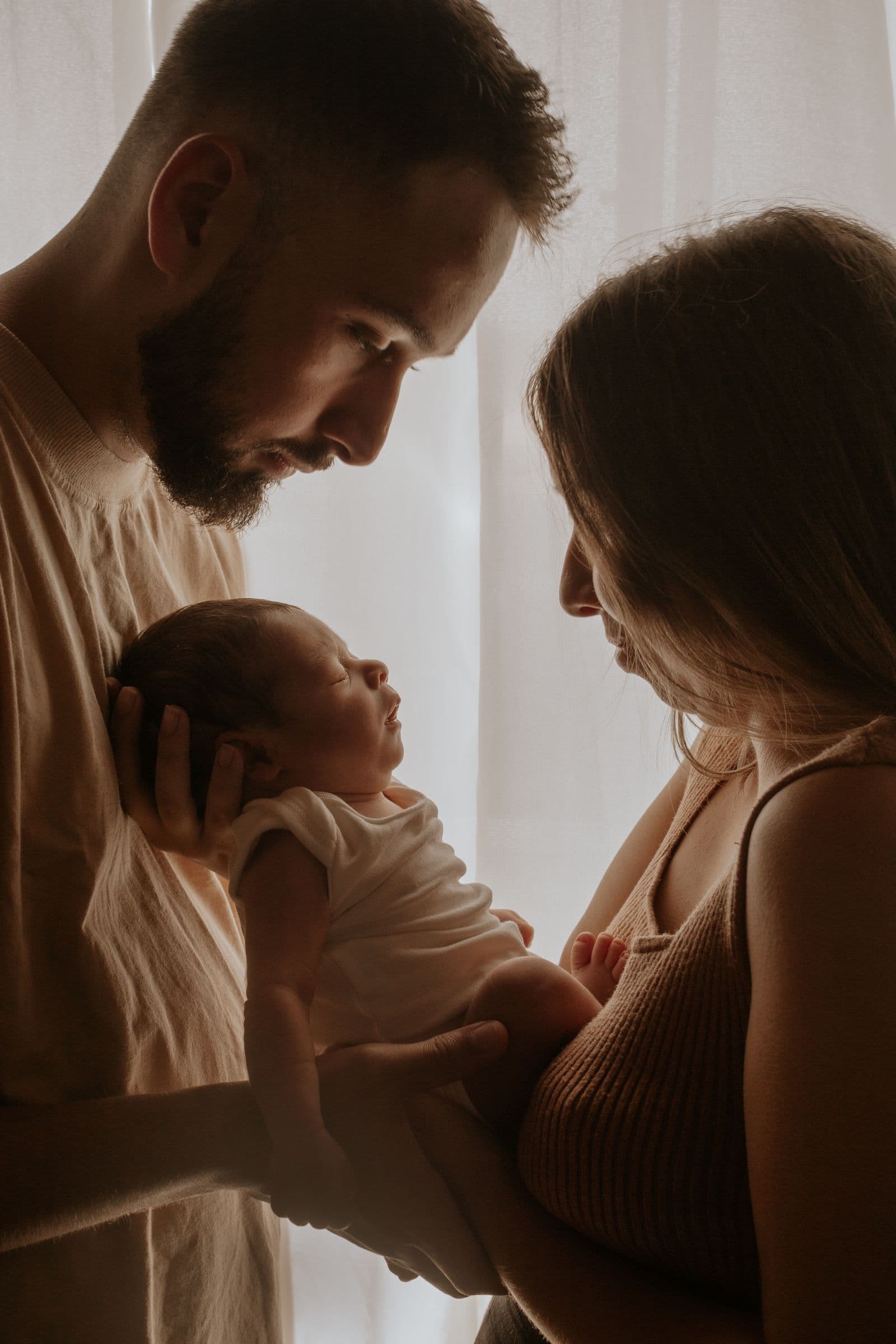 Two parents hold their newborn by a window in low warm light.