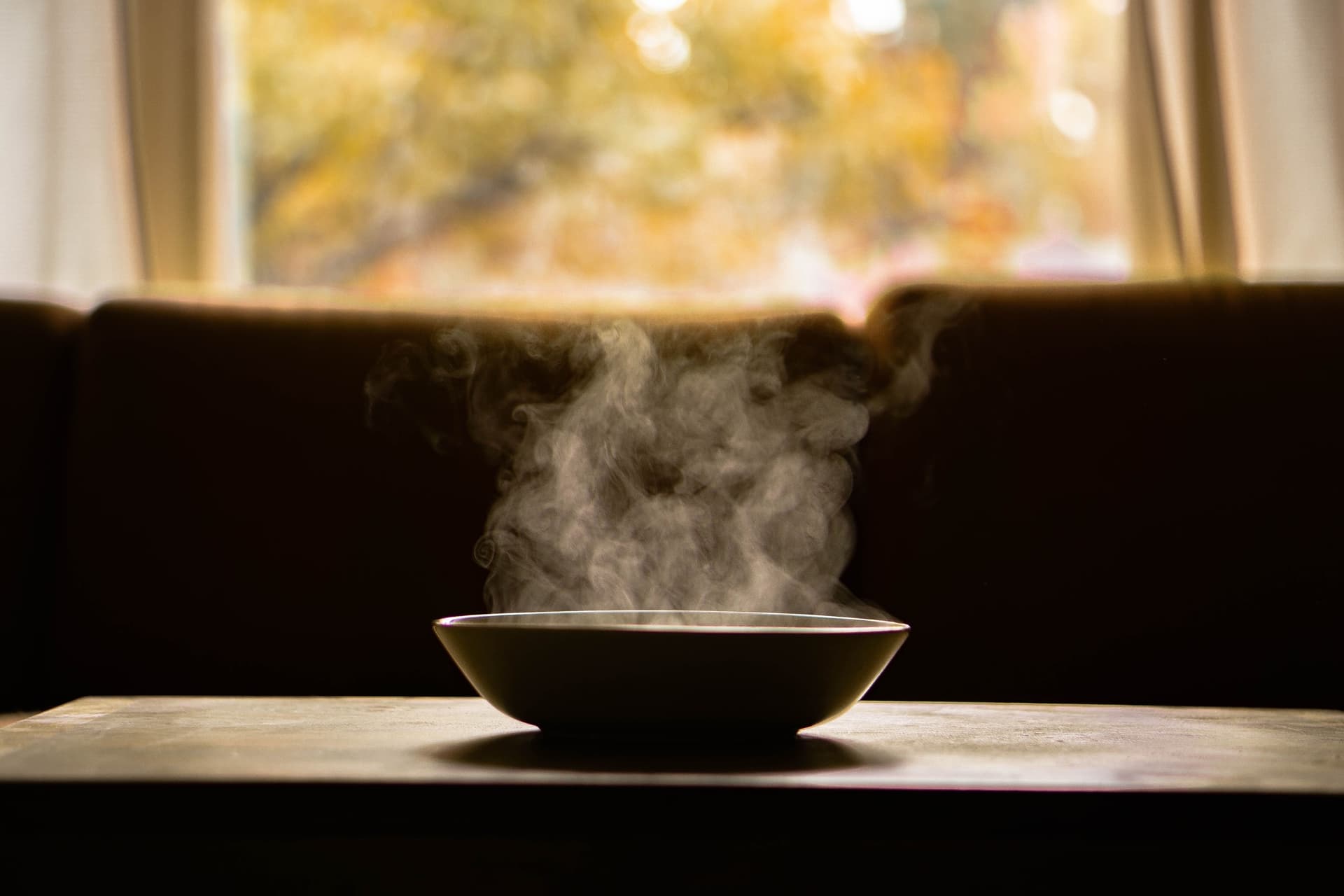 A bowl of soup steams on a wooden table in front of an autumn window.